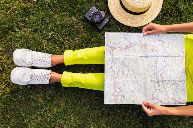 Photo gratuite vue de dessus des mains de femme noire tenant la carte, voyageur avec caméra s'amusant dans le style de mode d'été du parc, tenue de hipster coloré, assis sur l'herbe, pantalon jaune, jambes en chaussures de sport