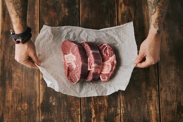 Vue de dessus des mains tatouées tiennent du papier kraft blanc avec trois grands steaks de jambe crus avec de l'os dessus, tout sur la vieille table en bois brossé