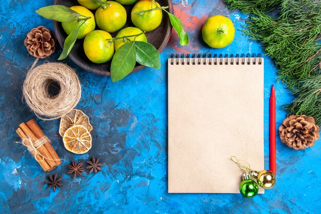 Vue de dessus mandarines jaunes avec des feuilles dans un bol en bois bloc-notes crayon rouge ornements de Noël bâtons de cannelle anis sur surface bleue