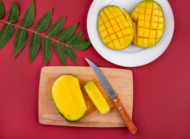 Vue de dessus de la mangue hachée avec couteau sur planche de cuisine en bois avec des tranches de mangue dans une assiette blanche avec des feuilles sur rouge