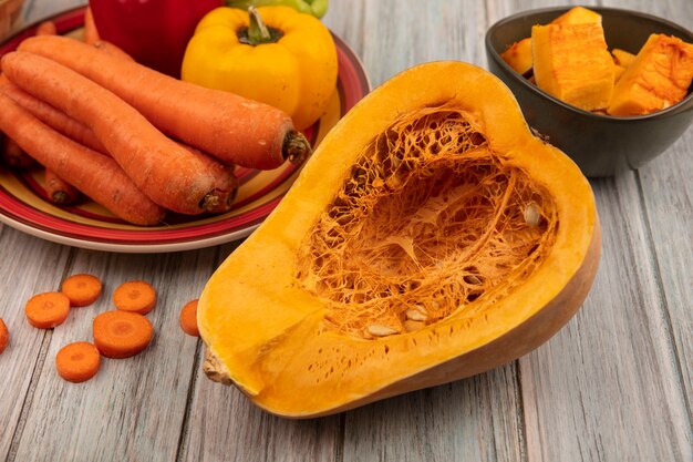 Vue de dessus de la moitié de la citrouille avec des poivrons colorés sur une assiette avec des carottes avec des tranches de citrouille sur un bol avec des carottes hachées isolé sur un fond en bois gris