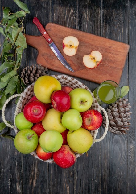 Vue de dessus de la moitié de la pomme et du couteau sur une planche à découper avec du jus de pomme panier de pommes pomme de pin et feuilles sur table en bois