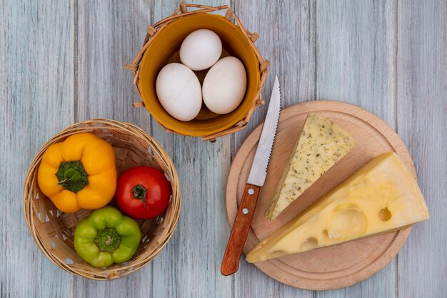 Vue de dessus des morceaux de fromage hollandais avec un couteau sur un support avec des poivrons et des œufs de poule sur un fond gris