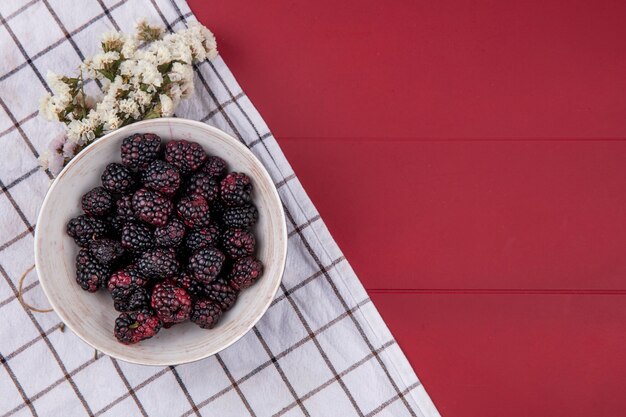 Photo gratuite vue de dessus de la mûre dans un bol avec une fleur blanche et une serviette à carreaux blancs sur une surface rouge