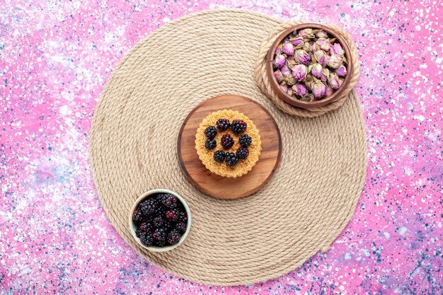 Vue de dessus des mûres fraîches à l'intérieur d'un petit pot blanc avec petit gâteau sur le bureau rose.