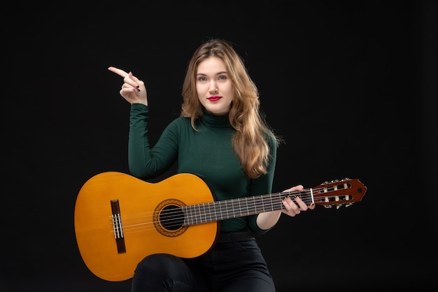 Vue de dessus d'une musicienne confiante tenant une guitare et pointant quelque chose sur le côté droit sur fond noir