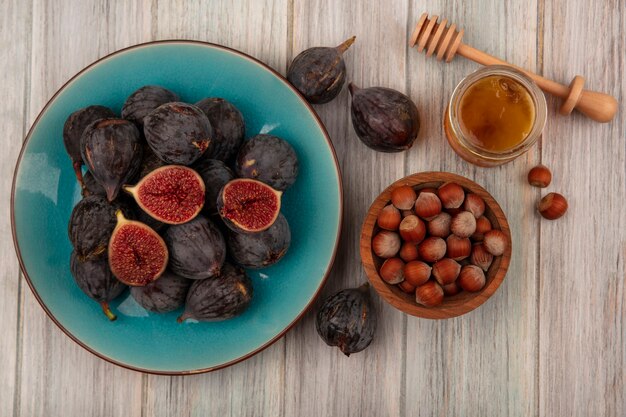 Vue de dessus des noisettes sur un bol en bois avec des figues de mission noires mûres sur un plat bleu avec du miel dans un bocal en verre et cuillère à miel sur un mur en bois gris