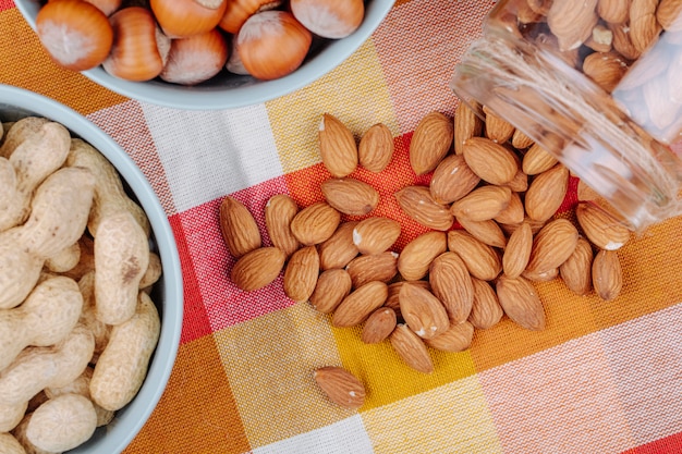 Vue de dessus des noix arachides noisettes dans des bols et des amandes dispersées dans un bocal en verre sur une serviette de table à carreaux