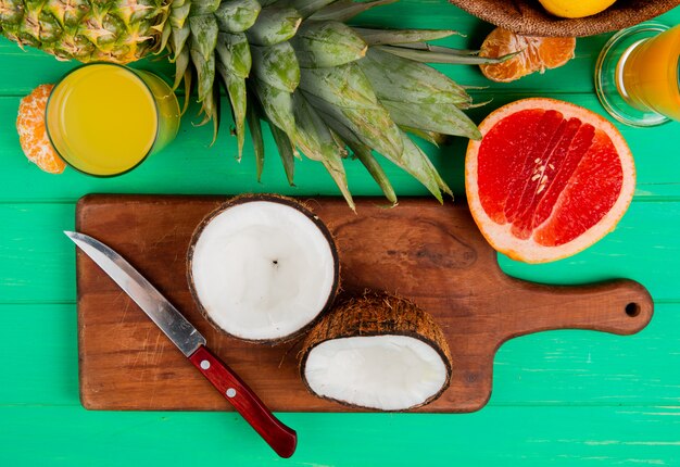 Vue de dessus de la noix de coco coupée à moitié et un couteau sur une planche à découper avec des pamplemousses ananas mandarine et jus d'orange sur fond vert