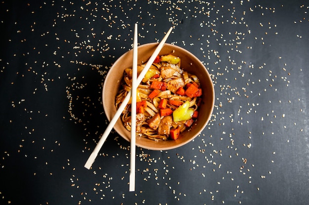 Photo gratuite vue de dessus des nouilles au poulet avec des légumes dans une assiette avec des baguettes et des graines de sésame sur fond noir