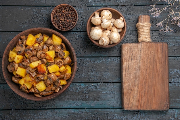 Vue de dessus de la nourriture dans un bol assiette de pommes de terre et de champignons à côté d'un bol de planche à découper au poivre noir bol de champignons blancs et de branches