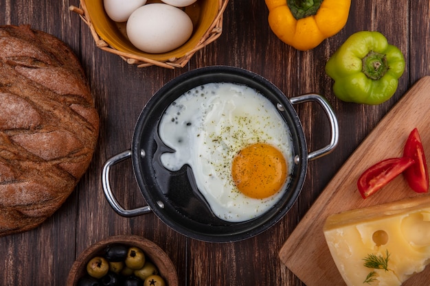 Vue de dessus des œufs au plat dans une poêle avec des olives œufs de poule dans un panier et poivrons tomate et litière maasdam sur fond de bois