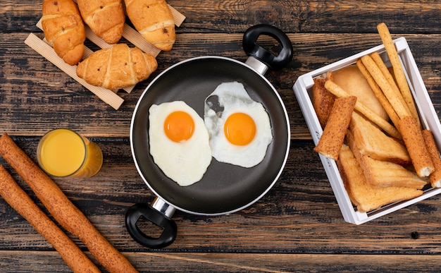 Vue de dessus des œufs au plat avec des toasts, des croissants et du jus sur une surface en bois sombre horizontal
