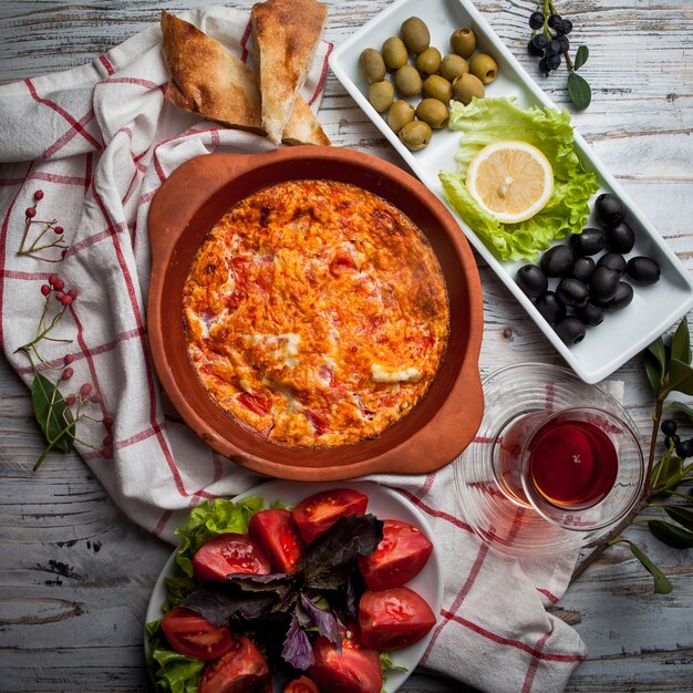 Vue de dessus des œufs au plat avec des tomates avec un verre de thé et un assortiment d'olives et de tomates dans une assiette en argile