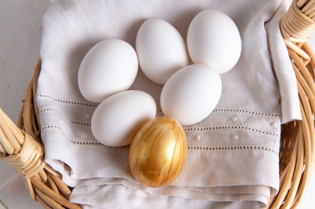 Vue de dessus des œufs entiers blancs à l'intérieur du panier avec des œufs d'or sur une surface claire
