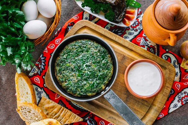 Vue de dessus des œufs fouettés kyukyu- aux herbes dans une casserole sur une planche avec du pain et du yaourt