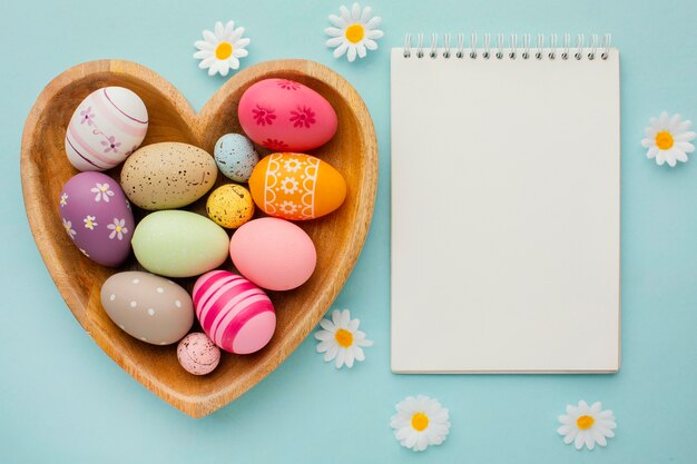 Vue de dessus des oeufs de Pâques colorés dans une assiette en forme de coeur avec cahier et fleurs