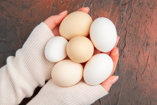 Vue de dessus des œufs de poule blancs à l'intérieur des mains des femmes sur une table sombre