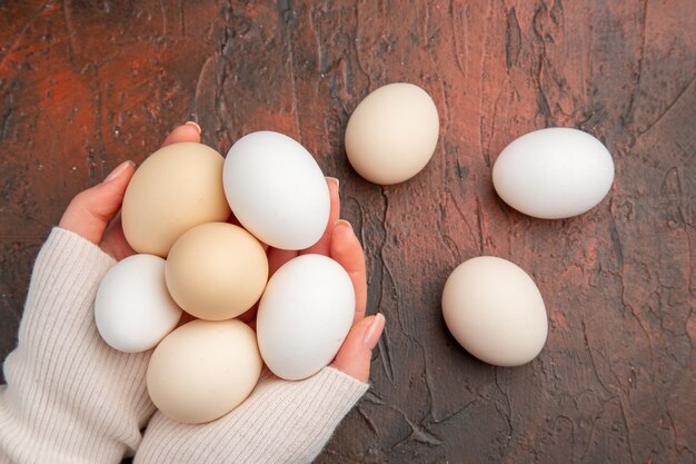 Vue de dessus des œufs de poule blancs à l'intérieur des mains des femmes sur une table sombre