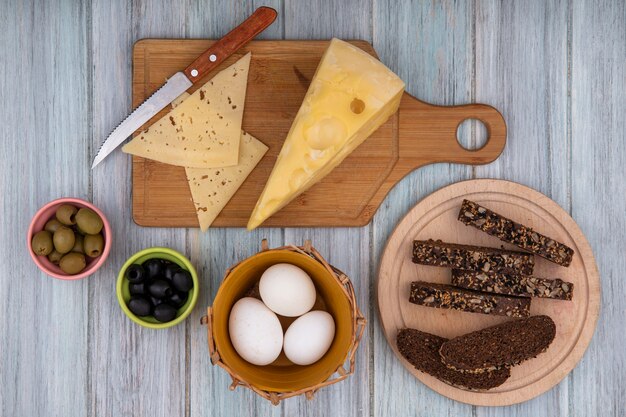 Vue de dessus des œufs de poule dans un panier avec des tranches de pain noir sur un support avec des fromages sur une planche à découper et un couteau sur fond gris