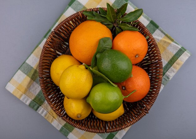 Vue de dessus des oranges aux citrons et limes dans un panier sur une serviette à carreaux jaune sur fond gris