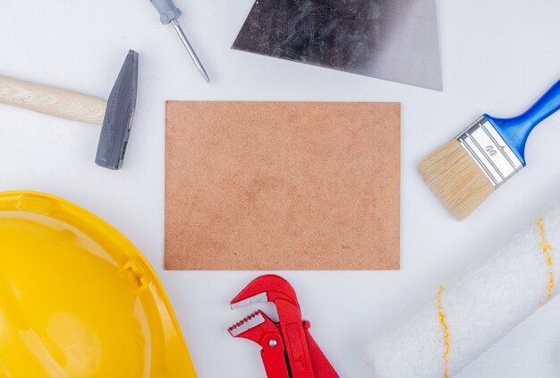 Vue de dessus des outils de construction comme brique marteau casque de sécurité tournevis clé à tube pinceau et couteau à mastic rouleau autour de tuile de mettlach sur fond blanc