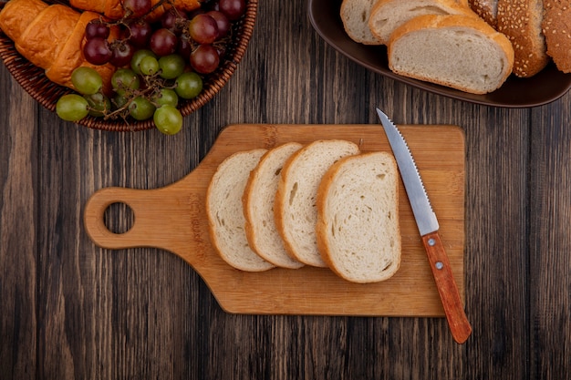 Vue de dessus des pains en tranches d'épis brun épépiné et blanc dans un bol et sur une planche à découper avec couteau et panier de raisin croissant sur fond de bois