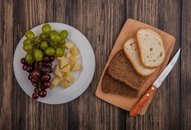 Vue De Dessus Des Pains En Tranches De Seigle Et Blancs Avec Un Couteau Sur Une Planche à Découper Et Une Assiette De Raisin Et De Fromage Sur Fond De Bois