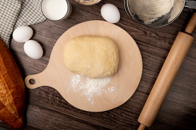 Vue de dessus de la pâte et de la farine sur une planche à découper avec du lait à oeufs rouleau à pâtisserie sur fond de bois