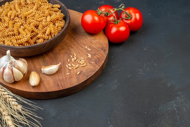 Vue de dessus des pâtes crues dans un bol brun riz à l'ail sur des pointes de planche de bois et des tomates avec des tiges sur fond noir