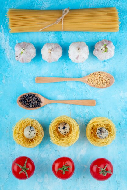 Vue de dessus des pâtes crues de différentes formes et types comme nid de pâtes spaghetti jaune avec de petits œufs de caille cuillère en bois avec des étoiles en forme d'ail pâtes et tomates fraîches sur fond bleu