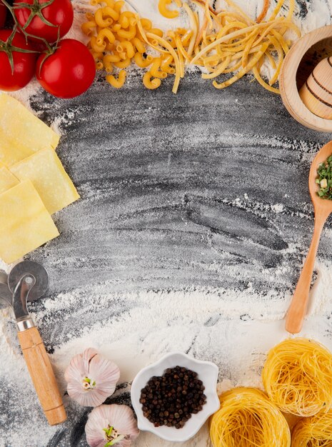 Vue de dessus des pâtes crues de différentes formes et types de tomates fraîches et d'ail sur fond noir avec de la farine avec copie espace