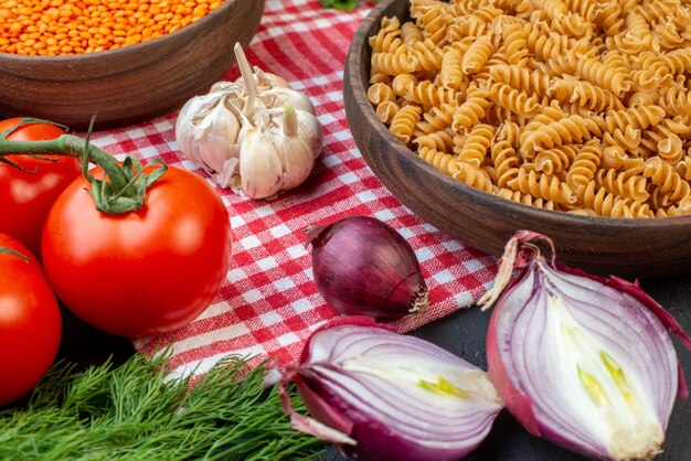 Vue de dessus des pâtes crues sur une planche à découper en bois sur une serviette dépouillée rouge et des légumes frais verts sur fond sombre