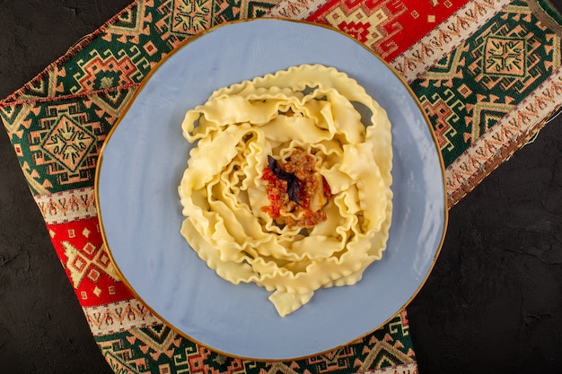 Une vue de dessus pâtes cuites savoureuses salées à l'intérieur de la plaque bleue ronde sur le tapis conçu et bureau sombre