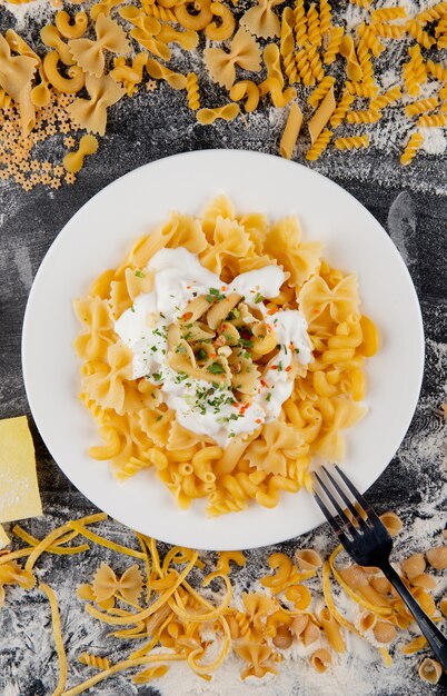 Vue de dessus de pâtes italiennes crues assorties et de pâtes farfalle cuites avec sauce à la crème dans une assiette blanche sur fond noir