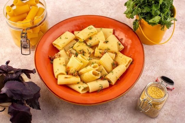 Une vue de dessus pâtes italiennes cuites savoureuses salées à l'intérieur de la plaque orange ronde avec des fleurs et des pâtes crues sur un bureau rose