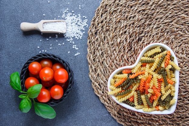 Vue de dessus des pâtes macaroni colorées dans un bol en forme de coeur avec des tomates, des feuilles, du sel de cristal sur un dessous de plat et une surface grise. horizontal