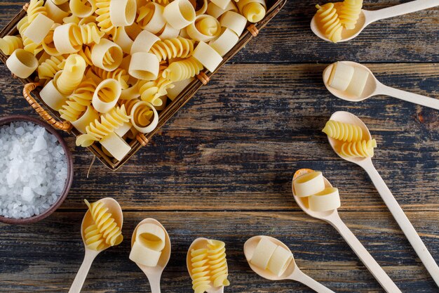 Vue de dessus des pâtes macaroni dans le panier et les cuillères avec du sel sur fond de bois. horizontal