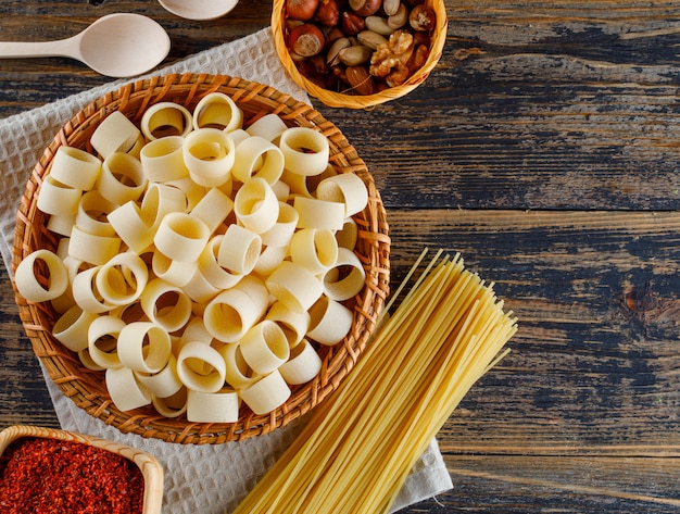 Vue de dessus des pâtes macaroni dans le panier avec des spaghettis, des cuillères, des noix diverses sur fond en bois. espace horizontal pour le texte