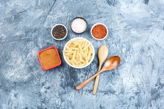 Vue de dessus des pâtes penne dans un bol blanc avec des cuillères en bois, des épices sur fond de plâtre gris. horizontal