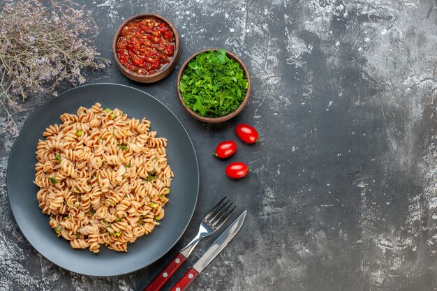 Vue de dessus pâtes rotini sur assiette ronde sauce tomate persil haché dans de petits bols tomates cerises fourchette et couteau sur l'espace libre de la table sombre