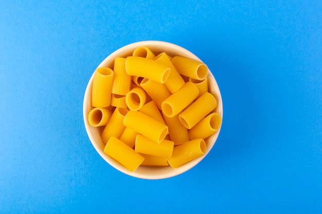 Une vue de dessus des pâtes sèches italiennes formées de petites pâtes crues jaunes à l'intérieur d'un bol rond de couleur crème isolé sur le fond bleu des pâtes alimentaires spaghetti italien