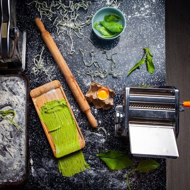 Vue de dessus des pâtes et des ustensiles de cuisine, y compris le rouleau avec des feuilles dans un bol sur fond texturé noir.