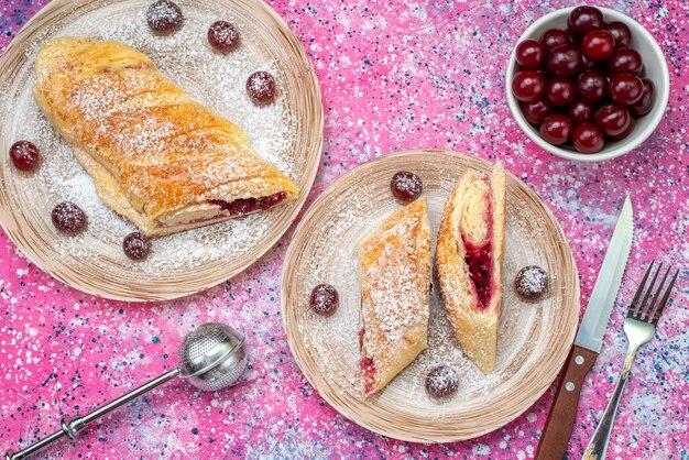 Vue de dessus de la pâtisserie aux cerises délicieuses et sucrées en tranches avec des cerises aigres fraîches à l'intérieur des assiettes sur le bureau de couleur biscuit sucré