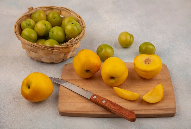 Vue de dessus des pêches jaunes fraîches sur une planche de cuisine en bois avec un couteau avec des prunes cerises vertes sur un seau sur un fond blanc