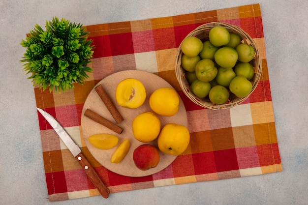 Vue de dessus des pêches jaunes sur une planche de cuisine en bois avec des bâtons de cannelle avec un couteau avec des prunes cerises vertes sur un seau sur une nappe à carreaux sur fond blanc