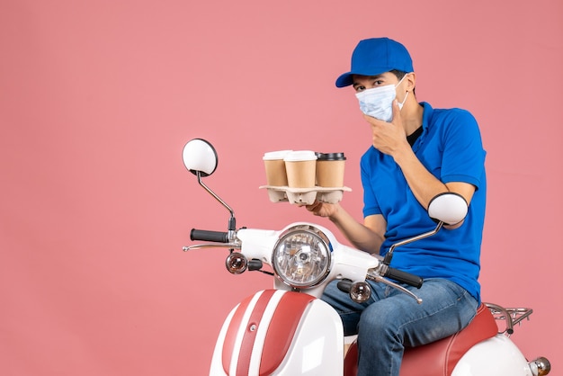 Vue de dessus de la pensée de l'homme de messagerie en masque portant un chapeau assis sur un scooter montrant des commandes sur une pêche pastel