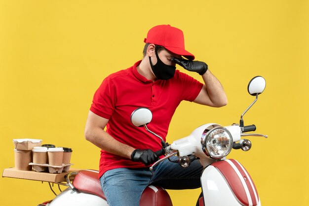 Vue de dessus de la pensée jeune adulte portant chemisier rouge et gants de chapeau dans un masque médical délivrant l'ordre assis sur un scooter sur un mur jaune