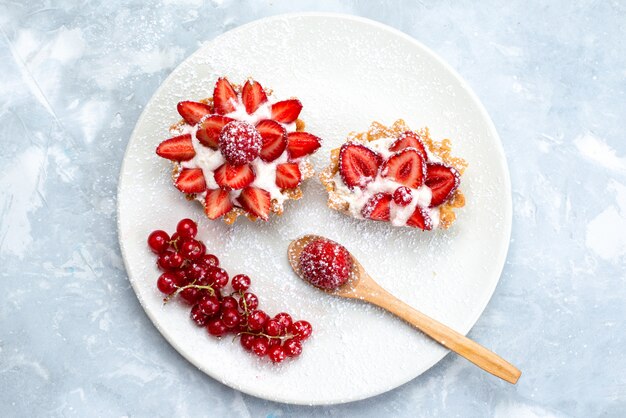 Une vue de dessus petit gâteau avec de la crème et des fraises fraîches tranchées à l'intérieur de la plaque blanche avec des canneberges rouges fraîches sur le bureau gris-bleu