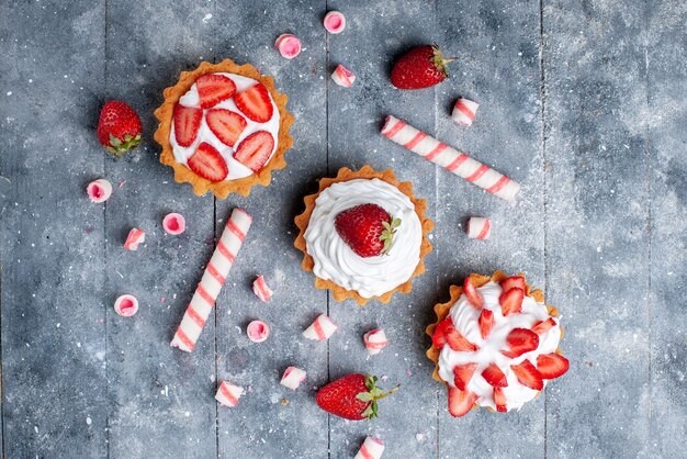 Vue de dessus petit gâteau crémeux avec des fraises fraîches et tranchées avec des bonbons de bâton sur le fond gris couleur douce de fruits photo cuire